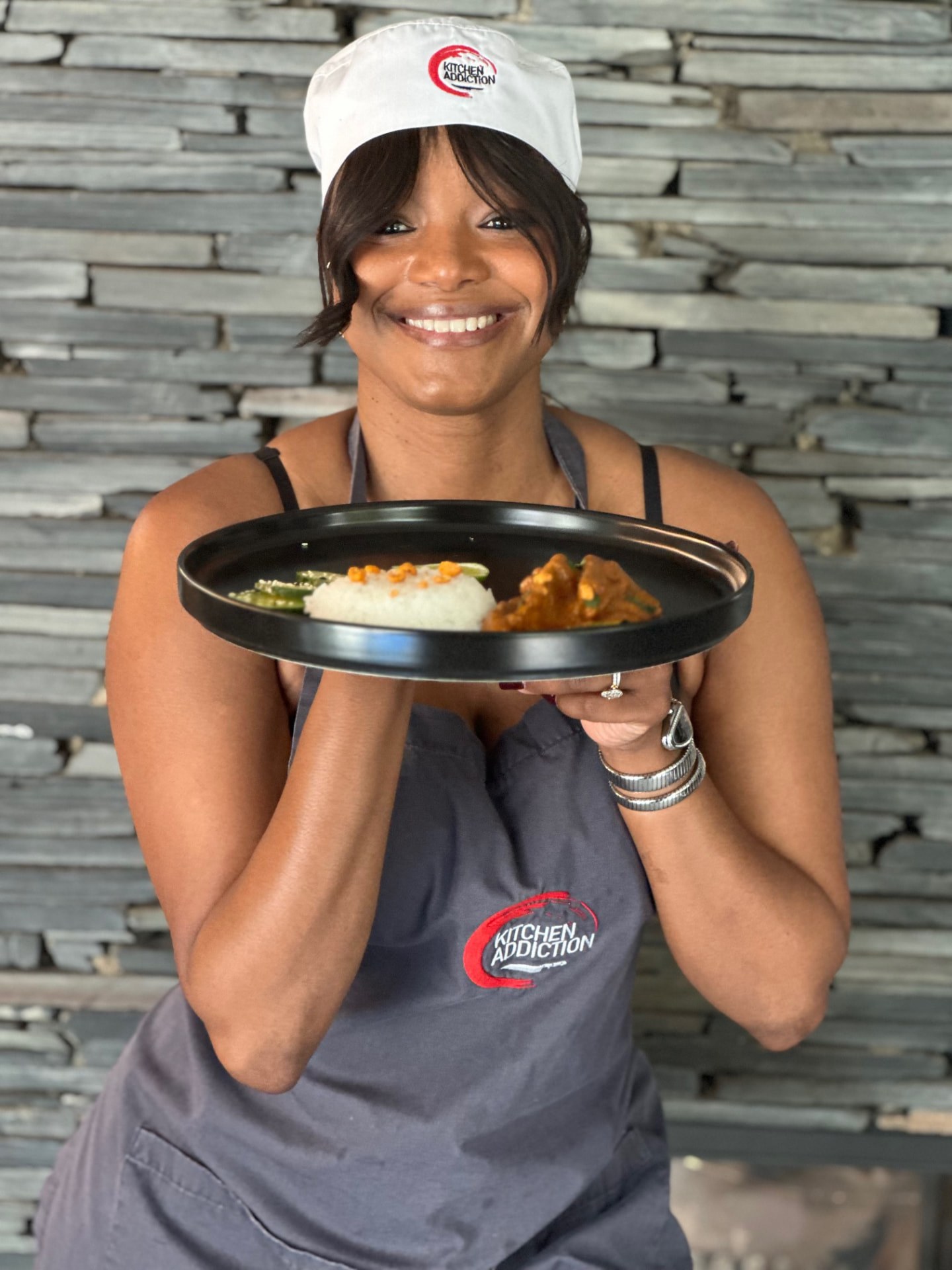 A guest in a Kitchen Addiction chef's hat smiling as she presents a plated curry with rice and cucumber salad